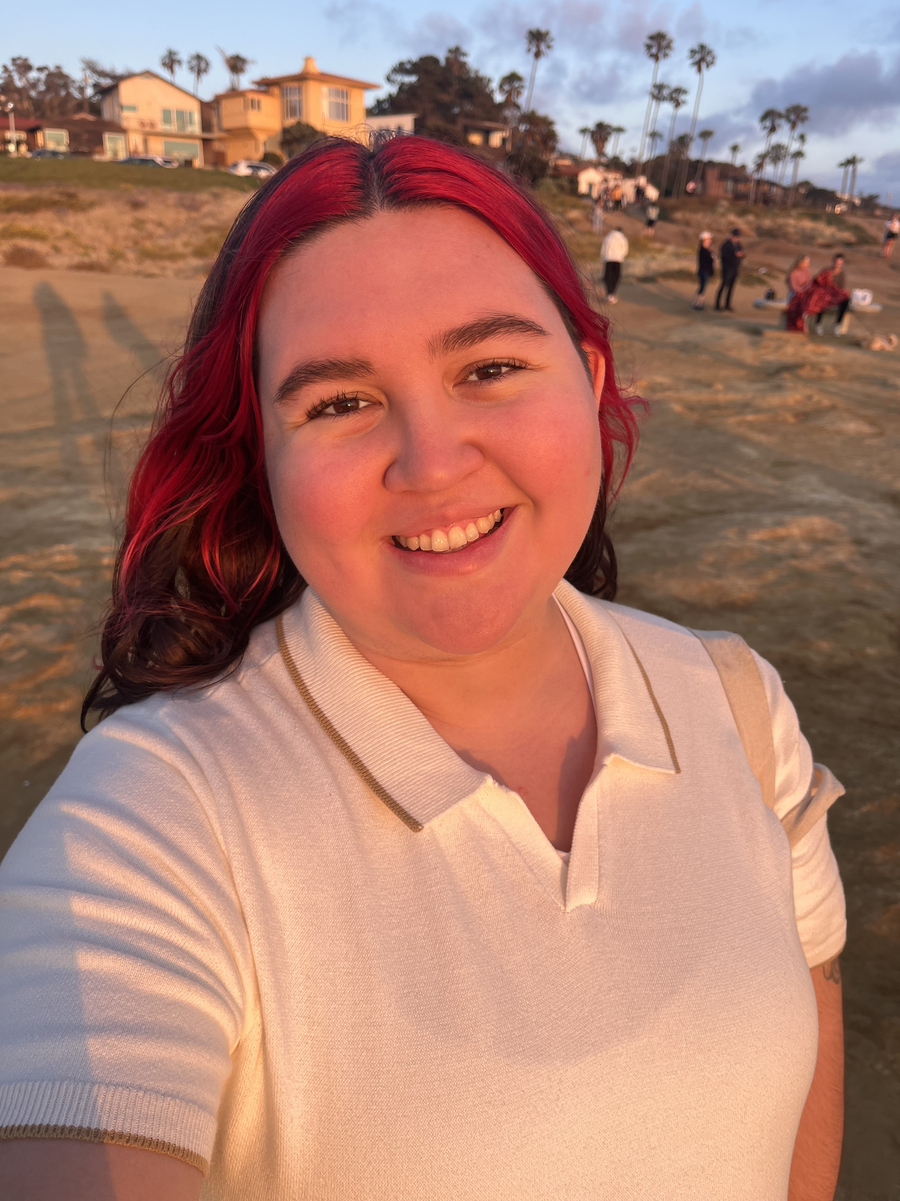 headshot of Latinx person at a beach, wearing a white button up shirt with dyed red hair.