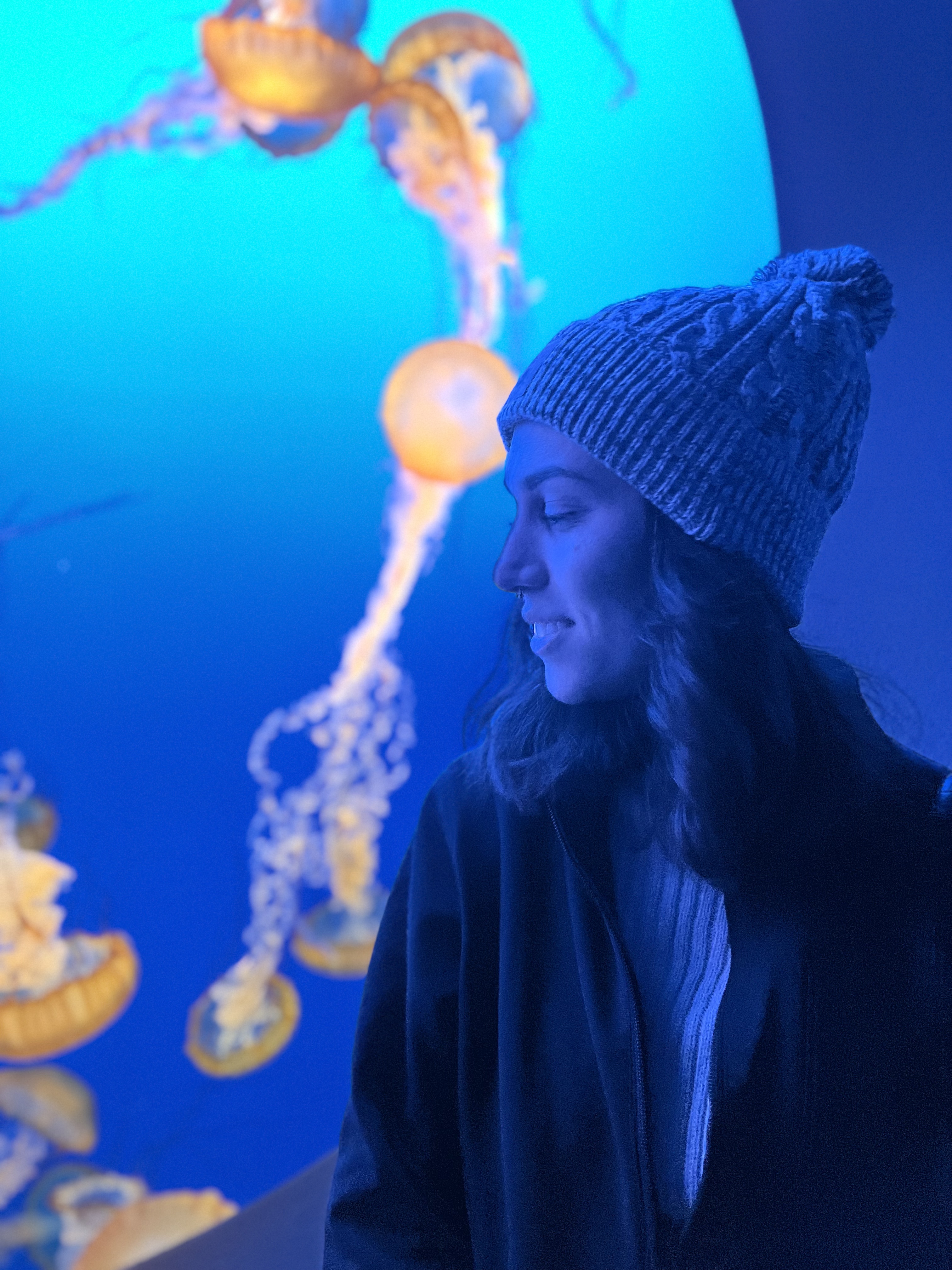 side profile of a white woman with long hair and a hat, standing in front of an aquarium exhibit in blue lighting